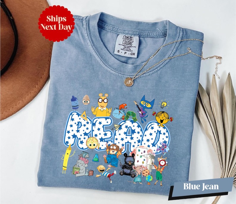 A blue t-shirt with a cartoon character design and the word "READ" printed on it, along with various other cartoon characters. The t-shirt is displayed on a white surface, and there is a brown hat and some decorative leaves in the background.