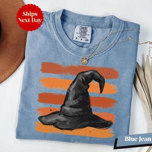 A blue denim t-shirt with a witch's hat design printed on it, along with a necklace and a brown hat.