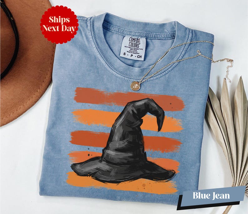 A blue denim t-shirt with a witch's hat design printed on it, along with a necklace and a brown hat.