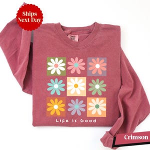 May include: Crimson long-sleeved shirt with a square floral design. The design features nine colorful flowers in square blocks, with the words "Life Is Good" below. The shirt is a solid crimson color.