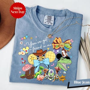 A blue denim t-shirt with a cartoon character design, including a dinosaur, a cowboy hat, and various other characters, along with the text "You've got me, you're a friend in me" and a "Ships Next Day" tag.