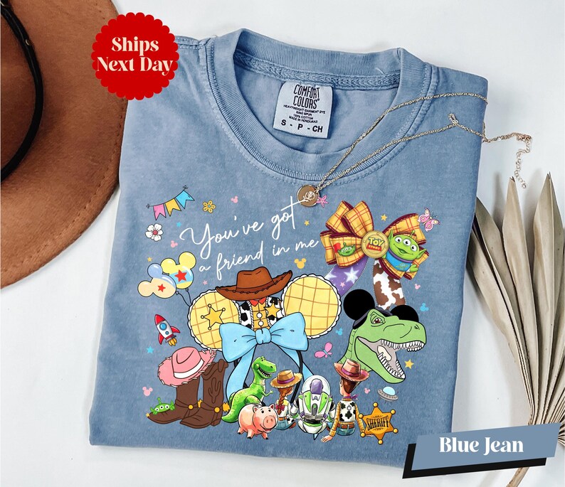 A blue denim t-shirt with a cartoon character design, including a dinosaur, a cowboy hat, and various other characters, along with the text "You've got me, you're a friend in me" and a "Ships Next Day" tag.