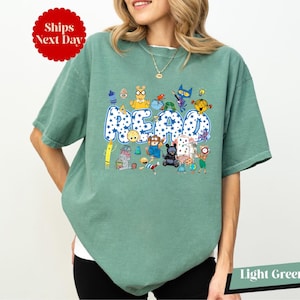 A woman wearing a green t-shirt with a colorful graphic design featuring various cartoon characters and the word "REPEAT" prominently displayed.