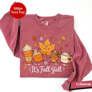 A pink long-sleeved t-shirt with a graphic design featuring cartoon characters and fall-themed elements such as leaves, a pumpkin, a cup of hot chocolate, and a football.