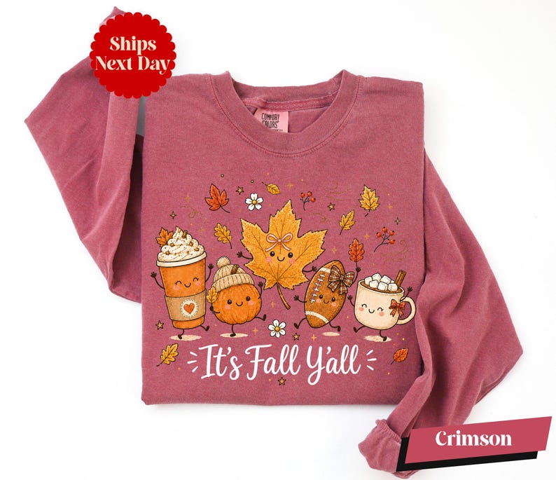 A pink long-sleeved t-shirt with a graphic design featuring cartoon characters and fall-themed elements such as leaves, a pumpkin, a cup of hot chocolate, and a football.