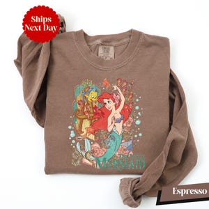 A brown t-shirt with a printed design of the mermaid character from the Disney animated film "The Little Mermaid".