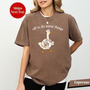 May include: An espresso-colored short-sleeve t-shirt featuring a graphic of a white goose dressed as a nurse, wearing a cap, stethoscope, and holding a clipboard and coffee cup. The text above the goose reads, off to do nurse things.