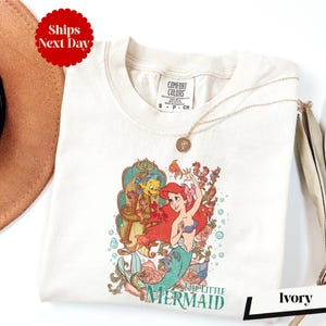 a white t-shirt with a colorful graphic of a mermaid character from the Disney film "The Little Mermaid". The t-shirt is displayed on a white background, along with a brown hat and a beige umbrella.