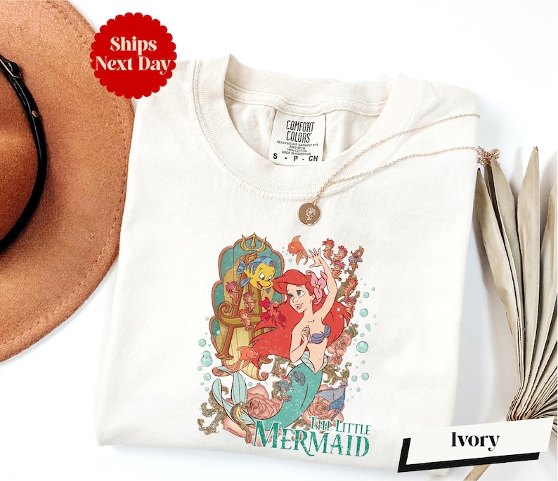 a white t-shirt with a colorful graphic of a mermaid character from the Disney film "The Little Mermaid". The t-shirt is displayed on a white background, along with a brown hat and a beige umbrella.