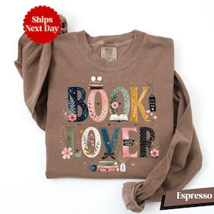 May include: An espresso-colored long-sleeve crewneck sweatshirt featuring the text BOOK LOVER in large, decorative letters. The letters are adorned with floral illustrations, small stacks of books, and tiny hearts. The text includes small labels reading book lover club and currently reading.