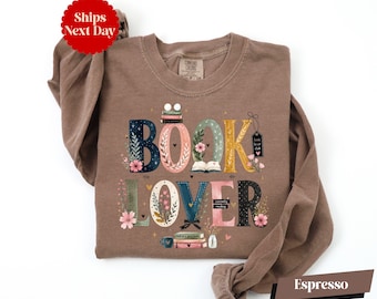 May include: An espresso-colored long-sleeve crewneck sweatshirt featuring the text BOOK LOVER in large, decorative letters. The letters are adorned with floral illustrations, small stacks of books, and tiny hearts. The text includes small labels reading book lover club and currently reading.