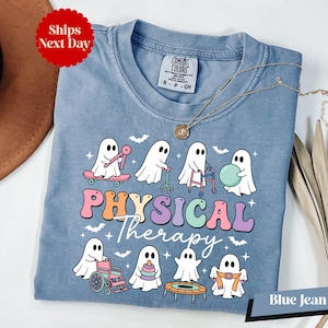 May include: A blue jean colored t-shirt featuring a graphic of white ghosts engaged in physical therapy activities. The text reads PHYSICAL Therapy in colorful, retro-style lettering. The ghosts are shown using a skateboard, walker, wheelchair, and exercise equipment, surrounded by small bats and stars.