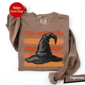 A brown t-shirt with a graphic of a witch's hat printed on it, along with a red circular sticker that says "Ships Next Day".