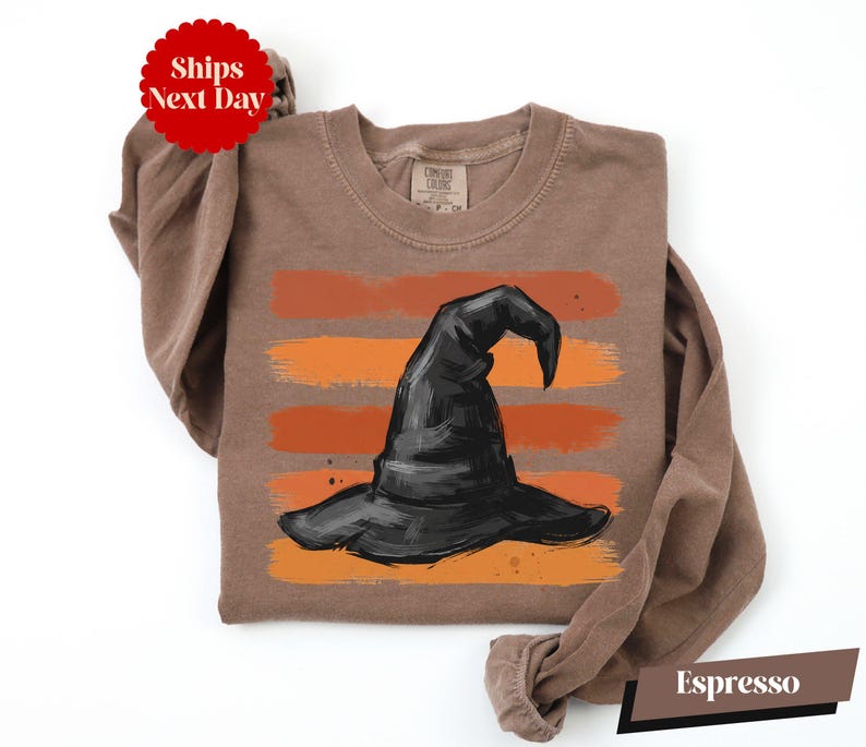 A brown t-shirt with a graphic of a witch's hat printed on it, along with a red circular sticker that says "Ships Next Day".
