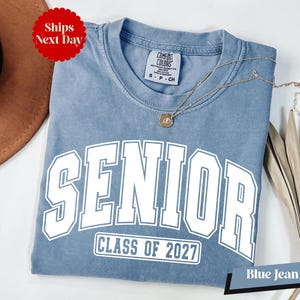 A blue t-shirt with the text "SENIOR CLASS OF 2027" printed on it, along with a necklace and a brown hat.