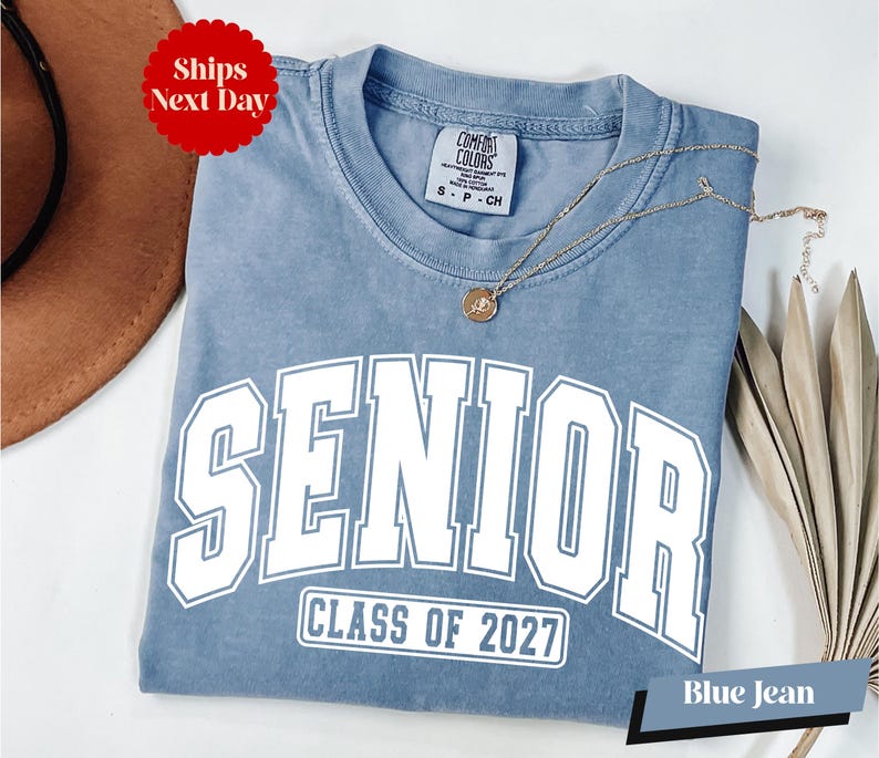 A blue t-shirt with the text "SENIOR CLASS OF 2027" printed on it, along with a necklace and a brown hat.