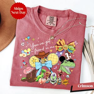A pink t-shirt with a cartoon character design, featuring various Disney characters such as Woody, Buzz Lightyear, and Timmy, along with other Disney-themed elements.