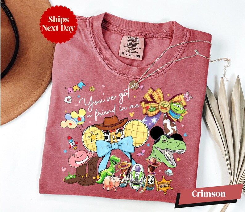 A pink t-shirt with a cartoon character design, featuring various Disney characters such as Woody, Buzz Lightyear, and Timmy, along with other Disney-themed elements.