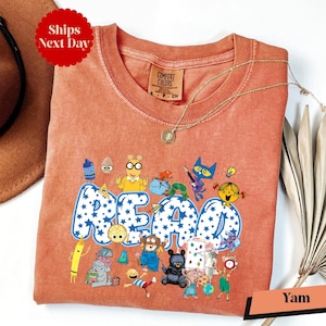 a pink t-shirt with a cartoon character design and the word "READ" printed on it. The t-shirt is displayed on a white background, along with a brown hat and a fan.