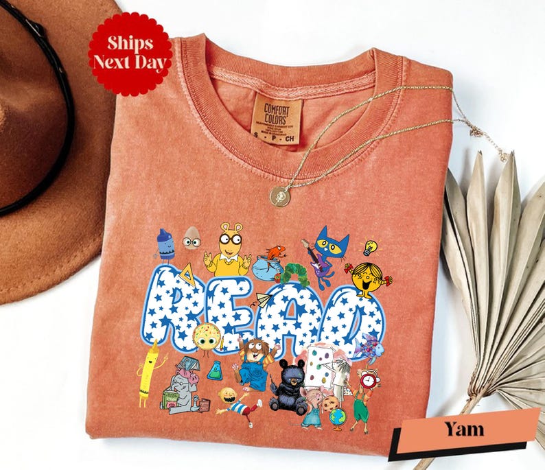 a pink t-shirt with a cartoon character design and the word "READ" printed on it. The t-shirt is displayed on a white background, along with a brown hat and a fan.