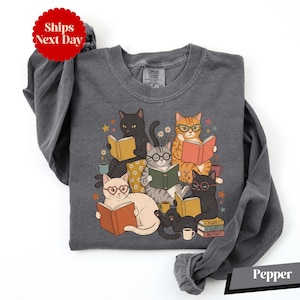 May include: A charcoal gray long-sleeve crewneck sweatshirt featuring a graphic illustration of five cats wearing glasses and reading books. The cats are surrounded by small stars, flowers, and coffee mugs. A stack of books reads Purr, Sleep, Repeat.
