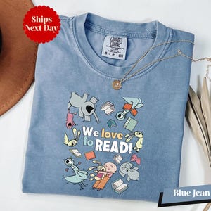 May include: A blue jean Comfort Colors t-shirt with the text "We love to READ!" and cartoon illustrations of animals and books. The shirt has a crew neck and short sleeves. A gold necklace is draped on the shirt.