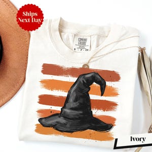 a white t-shirt with a black witch's hat printed on it, along with a brown hat and a necklace.