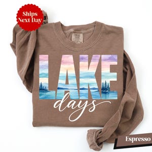 A brown long-sleeved shirt with the word "LAKES" printed on it, along with a watercolor landscape depicting a lake and mountains. The shirt is laid out on a white background, and there is a red circular sticker with the text "Shops Next Day" on it.