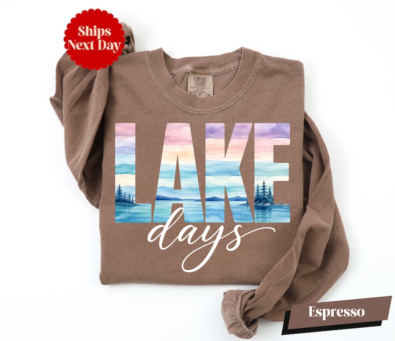 A brown long-sleeved shirt with the word "LAKES" printed on it, along with a watercolor landscape depicting a lake and mountains. The shirt is laid out on a white background, and there is a red circular sticker with the text "Shops Next Day" on it.