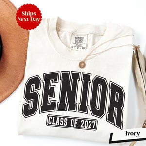 A white t-shirt with the text "SENIOR CLASS OF 2027" printed on it, along with a gold chain necklace. The t-shirt is placed on a white background, and there is a brown hat and a white card with the word "Ivy" written on it.