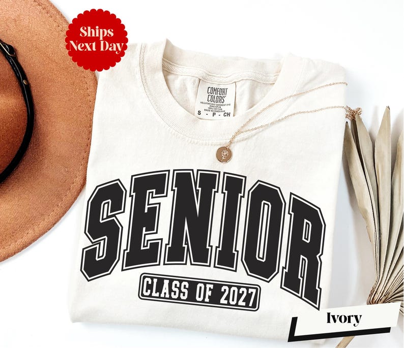 A white t-shirt with the text "SENIOR CLASS OF 2027" printed on it, along with a gold chain necklace. The t-shirt is placed on a white background, and there is a brown hat and a white card with the word "Ivy" written on it.