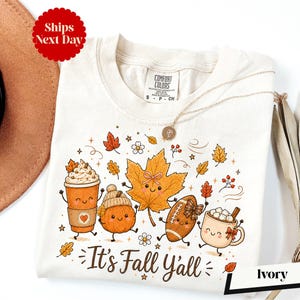 A white t-shirt with a graphic design featuring cartoon characters and fall-themed elements, such as pumpkins, leaves, and hot beverages.