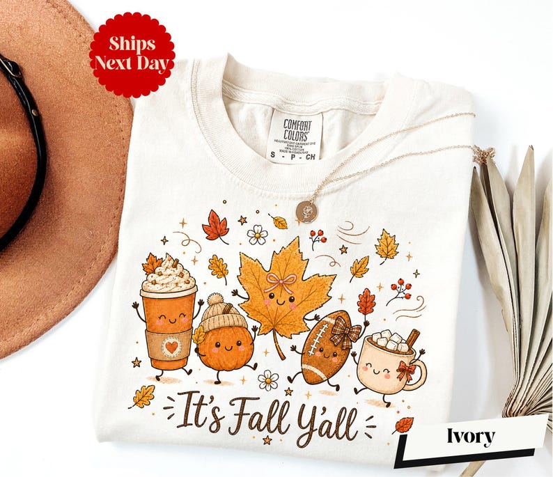 A white t-shirt with a graphic design featuring cartoon characters and fall-themed elements, such as pumpkins, leaves, and hot beverages.