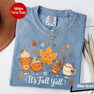 A blue t-shirt with a cartoon illustration of various fall-themed characters, including pumpkins, a coffee cup, and a football, along with the text "It's Fall Yall" and "Blue Jean".