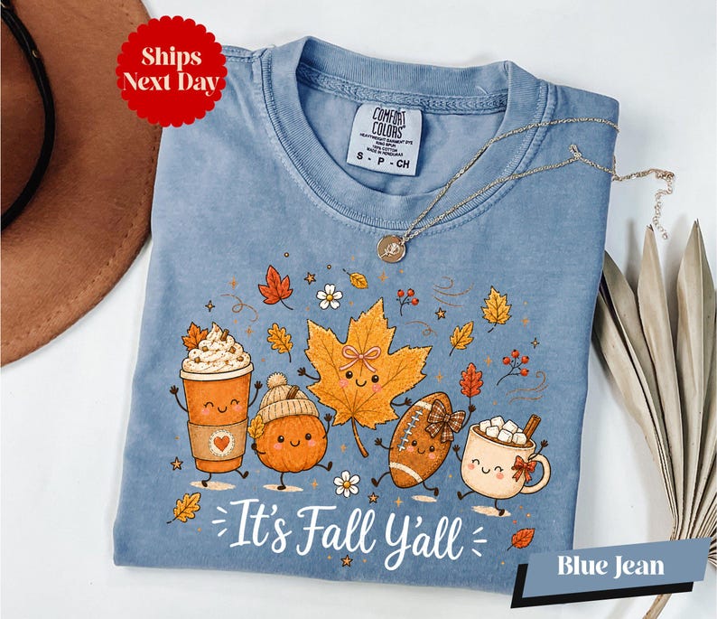 A blue t-shirt with a cartoon illustration of various fall-themed characters, including pumpkins, a coffee cup, and a football, along with the text "It's Fall Yall" and "Blue Jean".