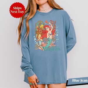 A woman wearing a blue long-sleeved shirt with a colorful graphic of a mermaid character from the Disney film "The Little Mermaid".