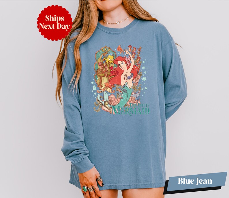 A woman wearing a blue long-sleeved shirt with a colorful graphic of a mermaid character from the Disney film "The Little Mermaid".
