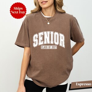 A woman wearing a brown t-shirt with the text "SENIOR CLASS OF 2027" printed on it.