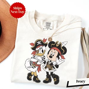 a white t-shirt with a cartoon image of Minnie Mouse and Donald Duck dressed as pirates, along with a pirate hat and a necklace. The t-shirt is displayed on a white background, and there is a brown hat and a white umbrella visible in the background.