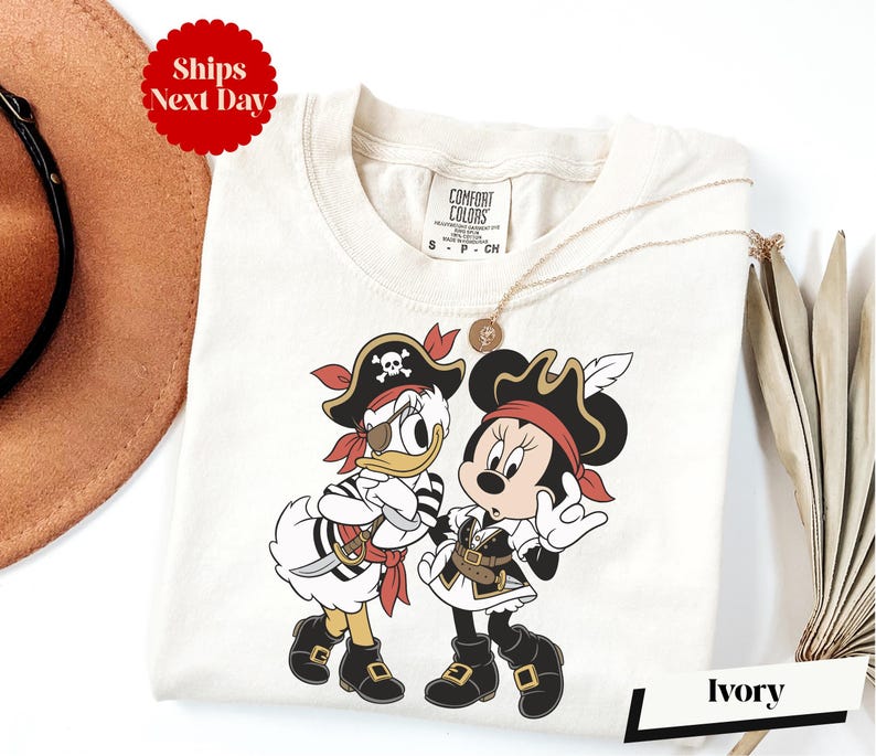 a white t-shirt with a cartoon image of Minnie Mouse and Donald Duck dressed as pirates, along with a pirate hat and a necklace. The t-shirt is displayed on a white background, and there is a brown hat and a white umbrella visible in the background.