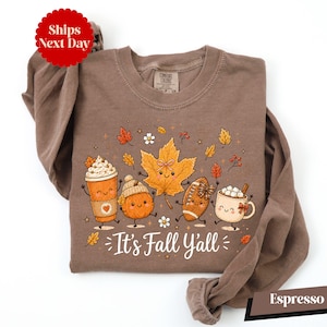 A brown long-sleeved t-shirt with a graphic design featuring cartoon characters and fall-themed elements such as leaves, pumpkins, and hot beverages.