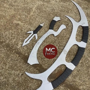 Könnte beinhalten: Ein Set aus drei silberfarbenen Klingen mit schwarzen Griffen, darunter ein Dolch, ein gebogenes Schwert und eine große sichelförmige Klinge. Die Klingen sind auf einer Betonoberfläche angeordnet. Das Logo "MC Knife Shop" ist sichtbar.