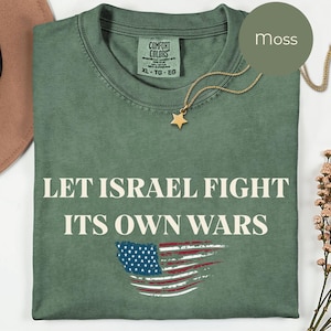 May include: A moss green t-shirt with the text "LET ISRAEL FIGHT ITS OWN WARS" and an American flag graphic. The shirt is folded and has a gold star pendant necklace. The tag reads "Comfort Colors".