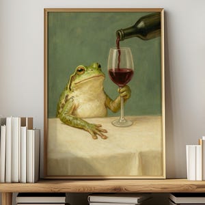 Pode incluir: Uma impressão artística emoldurada retrata um sapo segurando uma taça de vinho enquanto o vinho é derramado de uma garrafa. O sapo é verde e bege, contra um fundo verde. A mesa é coberta com uma toalha de mesa branca.