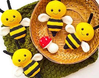 May include: Four handmade crochet bees with yellow and black striped bodies, white wings, and smiling faces. One bee is inside a woven basket with a red and white mushroom. The bees rest on a green crocheted surface.