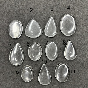 Può includere: Una collezione di undici cabochon di pietre preziose trasparenti e lucide in varie forme, tra cui ovali, a goccia e triangolari. Le pietre sono disposte su una superficie grigio scuro, ognuna con una forma unica e sottili motivi interni.