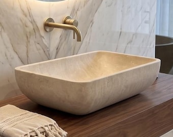 May include: A rectangular stone vessel sink sits on a thick wooden floating vanity shelf. A round backlit mirror is mounted above the sink with a brass wall-mounted faucet. Two amber glass soap dispensers and a beige fringed towel are on the shelf.