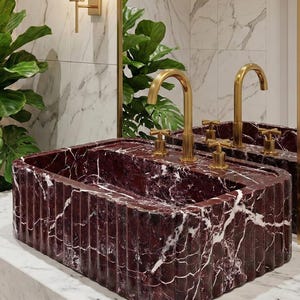 May include: A rectangular burgundy marble sink with white veining, featuring a fluted design. The sink has two gold-colored faucets and sits on a white marble countertop. The background includes a mirror, plants, and a wall-mounted light fixture.