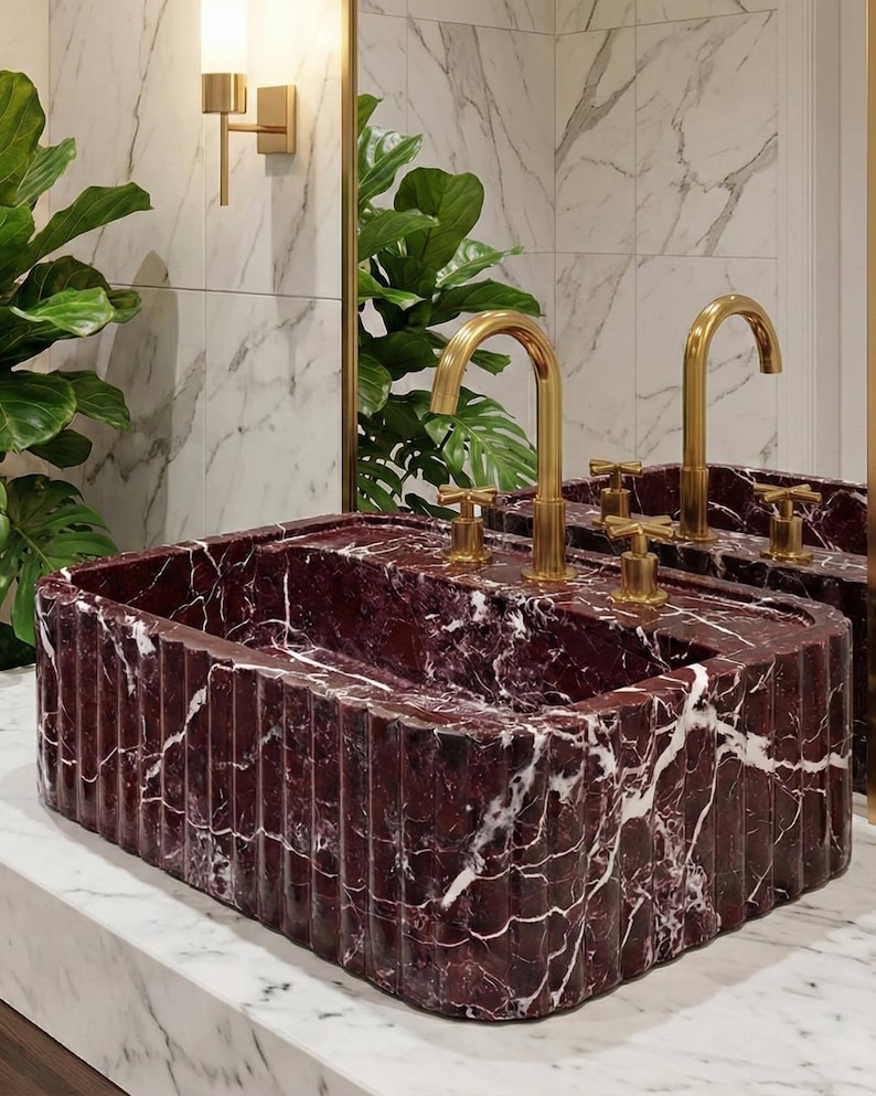 May include: A rectangular burgundy marble sink with white veining, featuring a fluted design. The sink has two gold-colored faucets and sits on a white marble countertop. The background includes a mirror, plants, and a wall-mounted light fixture.
