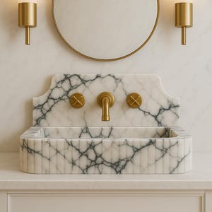 May include: A marble bathroom sink with gold fixtures, including a faucet and two sconces. The sink has a rectangular basin and a backsplash with a decorative edge. A round gold mirror hangs above the sink.